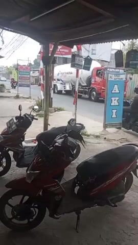 The video shows a street scene from under a shelter. In the foreground, two red motorcycles are parked on a table. In the background, a large white tanker truck labeled "PERTAMINA" is driving away from the camera, followed by a green dump truck. A person on a motorcycle is stopped near a sign that reads "AKI FZ BATTERY." The street is paved, and there are buildings and signs visible on the side of the road. The overall lighting suggests it is daytime, possibly overcast.