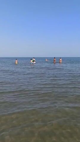 The video shows a clear blue sky above a calm sea with several people visible in the water. In the foreground, the water is shallow enough to see the sandy bottom. Further out, there are groups of people swimming and playing. One group appears to be gathered around a paddleboard. The water is transparent with gentle ripples. The horizon is flat and distinct between the sea and the sky.
