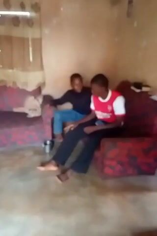 Two young males are in a room with two red sofas. The male on the left is wearing a black turtleneck and blue jeans. He is standing and leaning forward, appearing to touch or interact with the male on the right. The male on the right is seated on a sofa, wearing a red and white t-shirt and dark pants. He is looking towards the male on the left and holding a phone in his right hand. There are curtains in the background and the walls appear to be rough or textured. A metal cup is visible on the floor near the sofa on the left.