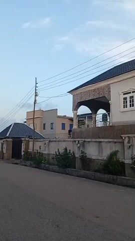 The video shows a street lined with houses on a cloudy day. The houses are of varying architectural styles, with some having modern designs and others appearing more traditional. There are trees and bushes along the street, and power lines are visible overhead. The street is paved, and there are no people or vehicles in the frame. The overall impression is one of a quiet residential area.