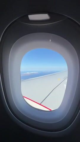 The video shows a view from an airplane window. The wing of the plane is visible, painted white with a red and white stripe featuring red arrows pointing downward. Beyond the wing, there is a clear blue sky and an ocean with landmasses that appear to be islands. The land is brown and rocky, and some clouds are hovering over it. The day is bright and sunny. The video zooms in slightly to focus on the islands.