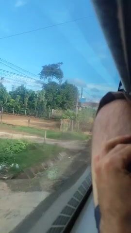 The video, filmed from inside a moving vehicle, shows a view through a window. The landscape outside consists of trees, houses, a dirt road, and vegetation. There are utility poles and wires visible. A person's arm and leg are partially visible in the foreground, resting against the window. The sky is blue with some clouds. The video appears to be taken in a rural or suburban area. There are signs with text in a non-English language.