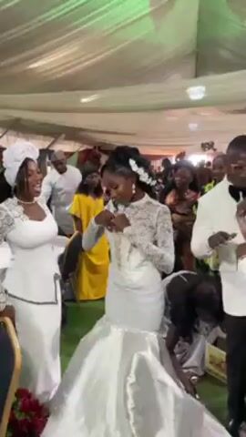 The video shows a wedding reception. A bride in a white mermaid-style wedding dress with lace sleeves and a fitted bodice is dancing with two other people. A woman in a white pantsuit and head covering dances to her left. To the bride’s right, a man in a white tuxedo with a black bow tie moves her dress train so that she can move freely. The reception venue is decorated with white fabric draping from the ceiling. Several guests are visible, wearing various formal attire. The bride appears to be of African descent.