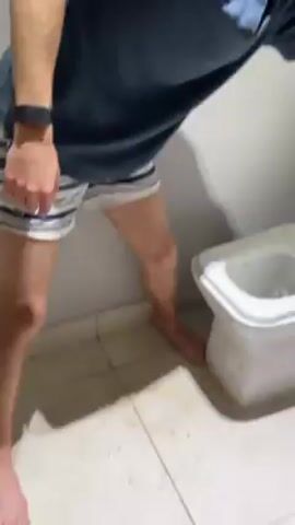A young person in a blue shirt and striped shorts leans over a white toilet. A second person, wearing a gray sweatshirt, stands nearby, holding a stack of white paper towels. The first person appears to be interacting with the toilet bowl. The scene takes place in a bathroom with white tile floors and walls. The first person has short blond hair and is wearing a black watch. The lighting in the bathroom is slightly dim. The focus of the video appears to be on the interaction with the toilet.