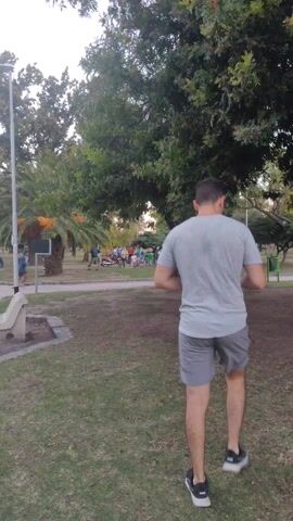 The scene is an outdoor park during daytime, with a group of people gathered on a grassy area near a pathway. The group consists of about a dozen individuals of varying ages, some standing and conversing, others seated in folding chairs. There is a motorcycle parked nearby, along with several bicycles. In the foreground, a man is seen running across the pathway, moving from left to right. The area is shaded by trees, including a palm tree, which provide partial cover. The overall atmosphere seems relaxed and casual.