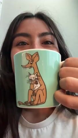A close-up shot focuses on a young woman holding a mug and bringing it to her lips to drink. The mug is light green and features a drawing of a brown, long-eared creature holding a white cat. The bottom of the mug is visible, displaying the text "ARABIA FINLAND" along with a small drawing of a Moomin character. The woman has dark hair and is wearing a white t-shirt. Her eyes are closed as she drinks, and she appears relaxed. The background is blurred, but hints at an indoor setting with a neutral-colored wall. The overall mood is peaceful and domestic. The timestamp indicates the scene is likely taking place in Lappeenranta, Finland.