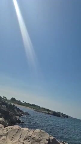 A bright sunbeam cuts across a clear blue sky over a rocky coastline and the ocean. The camera pans across the scene, showing the water and the horizon.