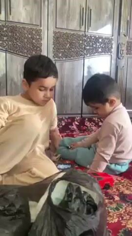 Two young children sit on a rug indoors in front of a wooden cabinet. The child on the left is wearing a beige outfit and looking down. The child on the right is wearing a light pink shirt and blue pants, also looking down. A red toy car is visible near the child on the right. In the foreground is a black trash bag. The patterned rug is predominantly red. The wooden cabinet behind the children has a decorative design.