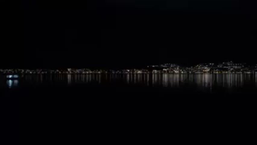 The video depicts a dark scene at night. A body of water dominates the foreground, its surface dark and reflective, mirroring the lights from the distant shoreline. On the horizon, a line of lights from a cityscape stretches across the frame, with varying intensities and colors that reflect on the water below. In the left of the frame, a single, dimly lit boat sits stationary on the water. The overall atmosphere is quiet and still.