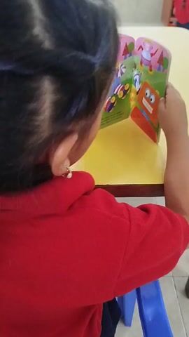 A young child with dark hair pulled back is sitting at a small, yellow table, intently looking at an open book. The child is wearing a bright red, long-sleeved shirt and small hoop earrings. The book features colorful cartoon illustrations of robots and other characters on a vibrant green and pink background. The child's hands are holding the book open, with one finger gently touching the page. The table is situated in a brightly lit room, and a blue chair is visible beneath it. Other individuals are present in the background, but their features are indistinct. The focus remains on the child and their engagement with the book.