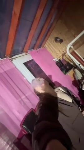 The video shows a person exhaling vapor. The environment appears to be a rustic room with wood-paneled walls and ceiling. Pink curtains are visible. In the second clip, the camera pans across a cluttered room with laundry on a rack and various items on shelves. A table with a green bowl and a white pitcher is also visible.