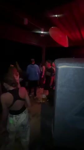 A group of people are dancing outdoors at night under changing colored lights. The lighting cycles through red, green, blue, and purple. There are trees visible in the background. One person in the foreground is wearing a black sports bra and patterned pants. In the background, several individuals are dressed casually. A large speaker is positioned in the right foreground. A red balloon is visible hanging from the ceiling.