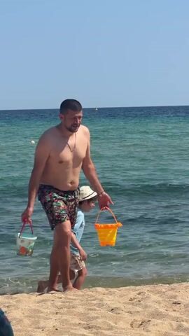 A man and a child are walking on a sandy beach near the water. The man is shirtless and wearing patterned swim trunks, carrying two buckets, one red and one orange. The child, wearing a blue shirt and patterned shorts, is slightly ahead of the man, also holding a bucket. They appear to be walking towards the water. The background shows the ocean with waves and a clear blue sky. Another man is visible further back in the water.