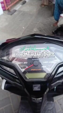 The video opens with a close-up of a motorcycle's dashboard, showing a speedometer and digital display. The perspective then shifts to behind the motorcycle, revealing a line of people on motorbikes waiting in traffic. The scene appears to be outdoors, with a covered walkway overhead and some red metal fencing visible on the left. The people are mostly facing away from the camera, and some are wearing casual clothing. The lighting suggests it is daytime. The video then continues to show the line of motorbikes moving slowly.