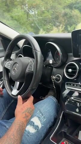 The video shows the interior of a car with a person's hands on the steering wheel. The person is wearing a denim jacket and has tattoos on their hand. The car is moving along a road surrounded by trees. The dashboard and center console are visible, including the car's infotainment system and air vents. A phone is mounted on the dashboard, and a car air freshener is hanging from the rearview mirror. The video captures a sense of movement and the car's environment.