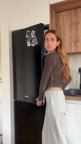 A woman opens a black refrigerator, grabs a bottle of ketchup, drinks some, closes the refrigerator door, and smiles. The woman has long brown hair, wears a brown long-sleeved crop top, and light grey sweatpants. The refrigerator is in a kitchen with white and brown wood cabinets.