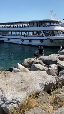 A medium shot captures three men fishing from a rocky shore, with a large, white passenger ferry docked behind them. The man on the left, wearing a black shirt and shorts, is actively casting his line into the water. The man in the middle, wearing a dark t-shirt and light pants, is looking down at the water, holding a fishing rod. The man on the right, wearing a light shirt, is seated on a rock, also fishing. The water is a clear, turquoise color, and the sky is bright blue. The ferry, named "ADA ISTANBUL" on its side, has multiple decks with people visible on the upper deck. The foreground is filled with large, gray rocks.