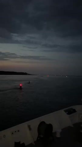The video shows the view from the bow of a ship at dusk. The sky is dark with dramatic clouds. The sea is calm and dark. In the distance, there are several navigation lights, indicating a channel or harbor entrance. A red buoy with a light is visible to the left, and a flagpole with a dark flag is in the center of the frame, on the ship's bow. The deck of the ship is visible in the foreground, with some equipment, including winches. The overall atmosphere is serene and somber.
