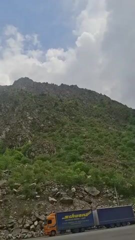 The video shows a road scene in a mountainous area. An orange and blue truck is parked on the side of the road, next to a rocky, green-covered hillside. The road stretches into a valley between the mountains. A white van and a white car are parked further up the road. A small group of people stands by a red umbrella, possibly at a roadside stall. The sky is partly cloudy, with a mix of sunlight and clouds. Power lines run alongside the road.