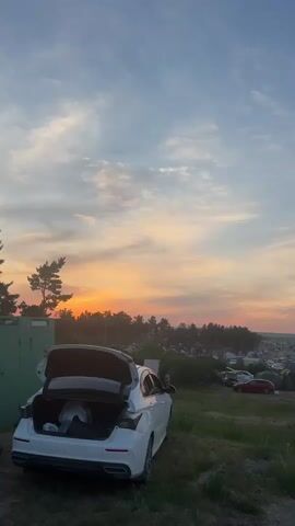 The video shows a campsite at sunset. In the foreground, a white car with its trunk open is parked on a grassy hill. Beyond the car, a dirt road winds through a field dotted with tents and other vehicles. The background features a treeline and a body of water. The sky is a mix of blue and orange, with scattered clouds. There are a few people visible in the distance, walking around the campsite. The overall impression is of a relaxed, outdoor gathering.