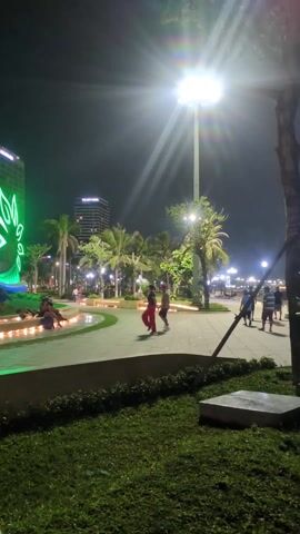 The video shows a well-lit park at night. Two people are dancing in the middle of a paved area. One person wears red pants and a dark shirt, while the other wears a patterned shirt, dark shorts, and a red hat. There are palm trees, benches, and a water feature lit with green lights in the background. Other people are walking or sitting nearby. A tall building and streetlights illuminate the scene.