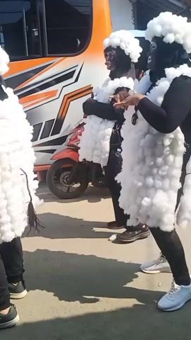 Three individuals, their faces painted black, are wearing costumes that resemble sheep. They are dressed in black clothing, and their costumes consist of white, fluffy material covering their torsos and heads, resembling wool. One individual is leaning forward, seemingly interacting with another, while the third stands slightly to the side. Behind them, a portion of an orange and white bus is visible, along with a red scooter parked in front of it. The ground appears to be a paved surface. The lighting suggests it is daytime.