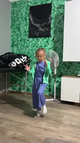 A young girl with blonde hair is standing in a room, wearing a blue jumpsuit and a light green cardigan. She is in the process of dancing or exercising, with her arms outstretched and her body in a dynamic pose. The background of the room features a vibrant green textured wall with a black framed picture hanging on it, a white fan, and a white cabinet. There is also a black bag on a foldable table to the left of the girl. The flooring is made of wood. The video is a short clip, likely a recording of the girl's movements.