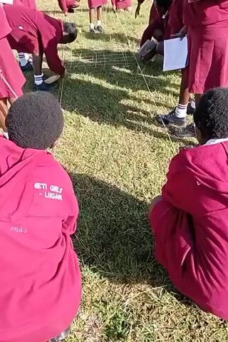 A group of young people wearing maroon skirts and maroon hooded sweatshirts, possibly uniforms, are gathered on a grassy lawn. They are working on a grid-like pattern marked out on the grass with string. Some of them are kneeling or crouching down, using tools that may be rulers or sticks, seemingly taking measurements or adjusting the string. They are also holding notebooks or clipboards with papers, perhaps recording information. The scene suggests a school activity or outdoor lesson, likely involving geometry or a similar practical exercise.