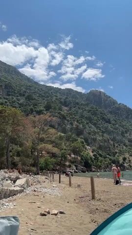 The video shows a view of a beach with a mountain backdrop under a partly cloudy sky. The beach is sandy with some wooden posts and rocky areas along the edge. A few people are visible on the beach, including a couple standing near the water's edge, as well as other individuals scattered along the shoreline. The water appears clear and calm. A tent, partially visible in the foreground, suggests someone is camping or spending the day at the beach. The mountain is covered in lush greenery and rocks, adding to the scenic landscape.