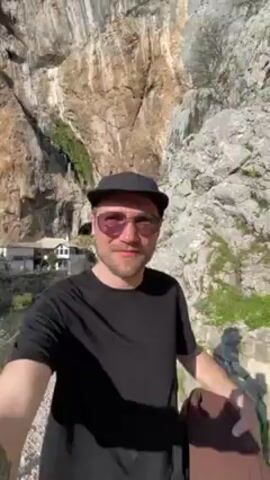 A man with sunglasses and a black baseball cap smiles into the camera, holding the camera out in front of him as he appears to walk along a path. A very tall cliff face is behind the man. In the distance are a group of buildings with stone roofs, appearing to be a village. A green river runs behind the buildings. The day is sunny.