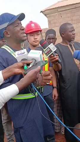 A group of men are gathered around someone giving an interview, holding microphones and recording with a phone. The man being interviewed is wearing a blue patterned outfit with a reflective vest, and a blue cap. Behind him, one man is wearing a red construction hat, and other men are wearing traditional Muslim attire. They are standing in what appears to be an outdoor construction zone with a building in the background. The microphones have logos, identifying KSMC and Arise News. The man being interviewed is gesturing with his hands while speaking.