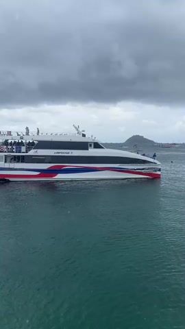 A static, medium shot captures a passenger ferry, "Lomprayah 7", sailing on a body of water. The ferry is white with red, white, and blue stripes along its hull. People are visible on the upper deck of the ferry. The sky is overcast with dark clouds. In the background, a small, hilly island is visible. The water is a dark green color.