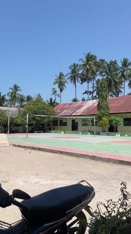 The video opens with a bright, sunny daytime view of a school or community courtyard in Matangpineung, Indonesia. In the foreground, part of a black motorcycle seat is visible. The courtyard features a multi-sport court with markings for basketball and what appears to be volleyball, complete with a net and a basketball hoop.

Beyond the court, several single-story buildings with red-tiled roofs are visible, surrounded by lush greenery and numerous tall palm trees against a clear blue sky. At 0:01, a man dressed in a black long-sleeved shirt and black trousers walks from left to right across the frame, carrying a white plastic bag in his left hand. He walks past a row of potted plants in the foreground.

The camera then pans slightly to the right, focusing on the potted plants in black containers arranged on a stand. In the background, the school buildings remain visible, and several people, likely students, can be seen gathered near the building entrances. The scene depicts a serene and sunny environment, typical of a school campus in a tropical region. No discernible text is visible.