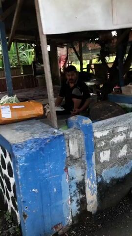 A man is sitting in a small, open-air stall. He is wearing a black t-shirt and has dark hair and a mustache. He is looking down, and his hands are not visible. To his left, there is a large, brown package wrapped in plastic and tape, with a white shipping label. Behind the man, there are some indistinct objects in the stall, including what appears to be a large, round, dark-colored object, possibly a turtle or a large fruit. The stall itself is made of wood and appears to be somewhat weathered. In front of the stall, there is a low wall, partially painted blue and white, with some visible graffiti or wear. The environment appears to be outdoors, with some greenery visible in the background, suggesting it might be a rural or semi-rural setting.