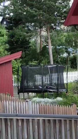 A person is jumping on a trampoline in a backyard. The trampoline is enclosed by a safety net. There is a red shed to the left of the trampoline, and a red house to the right. A wooden fence is in the foreground. The weather is overcast, and there are trees and bushes in the background.