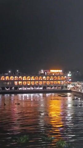 A night time wide shot of a two-story building on the water. The building has several illuminated arched windows on each floor. The name of the building is displayed in glowing letters on the roofline. The water is reflecting the light from the building. There are some plants floating in the water. A walkway extends from the building and people can be seen walking on it.