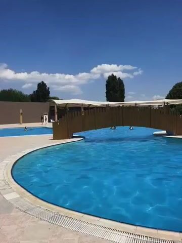 The video shows a swimming pool on a sunny day. A wooden bridge spans across a section of the pool, and there are people swimming and relaxing. The water is a bright blue, and the sky is clear with a few white clouds. There are trees and buildings in the background, suggesting a resort or a water park. The camera pans slightly, giving a view of the entire scene.