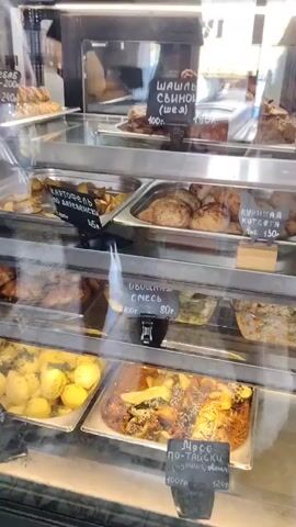 The video shows the inside of a food display case filled with various dishes. The display case appears to be in a cafeteria or a similar establishment. The dishes are arranged in metal trays. On the top shelf, there are roasted meats, possibly chicken and pork. There are also signs with prices and descriptions in Russian, such as "шашлык свиной (шея) 190 руб." and "курица котлета 130 руб." On the middle shelf, there is a dish of roasted potatoes with herbs, labeled "картофель по деревенски 45 руб." and another dish of mixed vegetables, labeled "овощная смесь 80 руб." The bottom shelf contains more roasted potatoes and another dish with meat and vegetables, labeled "мясо по-тайски 120 руб." The lighting is bright, and the food looks freshly prepared. The camera is moving slightly, giving a panning view of the display case. There are no people visible in the video.