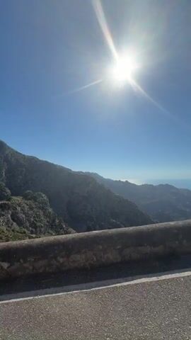 The video shows a sunny day overlooking a mountain range with the sea in the distance. The sun is bright in a clear blue sky.  A low stone wall runs along the foreground, next to a paved road. A woman leans into the frame momentarily, smiling and squinting into the sun, before disappearing.