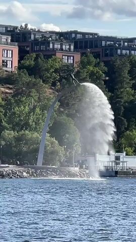 A large water fountain erupts from a structure at the edge of a body of water. A statue of a person is visible on the structure, appearing to be in a diving pose with water spraying from their hands. In the background, there are trees and modern buildings on a hill overlooking the water. The sky is blue with scattered white clouds. The water in the foreground is choppy.
