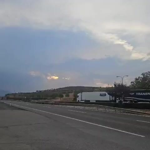 A truck drives by on a highway next to a rural landscape of hills and trees under a cloudy sky.