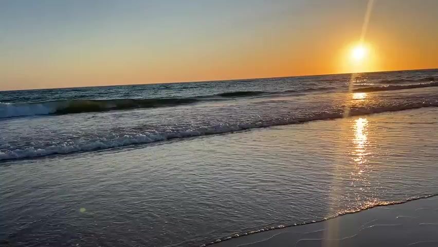The video captures a sunset over the ocean from a beach. The sun is a bright orb setting on the horizon, casting a golden-orange glow across the sky and reflecting on the water. Gentle waves are rolling onto the sandy shore. The sand is wet and reflective where the water has receded. The water is a deep blue-green, and the sky transitions from a clear blue at the top to a warm orange near the horizon. There are no people or man-made objects visible in the frame. The scene is serene and natural, focused on the beauty of the coastal environment at dusk.