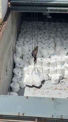 The video shows the interior of a cargo hold on a ship, filled with numerous large white sacks. A crane, suspended from above, is in the process of lifting a bundle of four smaller white sacks. These sacks are hanging from the main crane hook by ropes. The cargo hold is packed tightly with rows upon rows of the larger white sacks, creating a densely packed environment. The sacks appear to be made of a durable material, possibly woven polypropylene, and are sealed at the top. Some of the larger sacks have blue markings or labels visible. The floor of the cargo hold is made of metal grating or plating.

The overall scene suggests a loading or unloading operation is underway. The lighting indicates it is daytime, and the weather appears clear, as the sunlight is strong enough to create shadows. The context provided states the location is Uribia, Colombia, which is a port city. The activity is clearly the movement of bulk goods, likely being transferred onto or off the ship. There are no people visible in the frame.

Notable details include the scale of the operation, with a vast quantity of goods stored in the hold. The crane and its rigging are a focal point, highlighting the mechanism used for moving the cargo. Visible text on the ship's structure includes "19.00T" which likely indicates a weight measurement, possibly the capacity or current load. The repetition of the white sacks emphasizes the uniformity and volume of the cargo. The emotions conveyed are primarily industrial and functional, focusing on the logistics of shipping and trade.