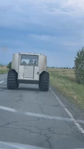 An unusual, boxy, light-colored vehicle with extremely large, thick tires is driving away from the camera on a cracked asphalt road. The vehicle has tinted windows, and several silhouettes of people can be seen inside. The vehicle appears to be on a paved road in a rural setting, with fields of grass and some trees visible on either side. The sky is cloudy.