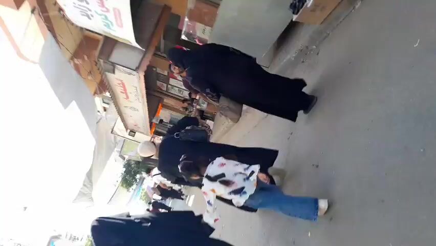 This video captures a street scene, likely in Deir el-Balah, Palestinian Territories. The perspective is from someone walking, looking down and to the right. Several people are visible, mostly women in dark abayas and hijabs, suggesting a modest dress code. One person in the foreground is wearing a light-colored patterned top and jeans, and another person appears to be a child walking with others.

The setting is an outdoor marketplace or street lined with shops. A large white awning or canopy is prominent on the left side of the frame, providing shade. Shops with displays and signage are visible along the street. One sign, partially obscured and tilted, has Arabic script that appears to include "ايس كريم" (ice cream). Another sign above a shop also contains Arabic writing. Boxes are stacked near a shop on the right.

The activity appears to be general pedestrian traffic, with people walking along the street, possibly shopping or commuting. The lighting suggests it is daytime, possibly late morning or afternoon, with bright sunlight creating glare and lens flare in some parts of the video. The overall mood seems ordinary, depicting everyday life in this location. The presence of a market suggests it could be a busy commercial area.