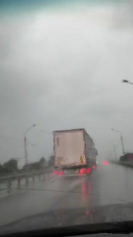 The video captures a view through a windshield during rainfall. The windshield wipers are intermittently clearing the view, showing a highway scene. A white truck with red taillights is visible ahead, driving on the wet road. Other vehicles with their taillights illuminated are also present. Streetlights line the side of the highway, and the sky is overcast, indicating poor weather conditions. The perspective is from inside a moving vehicle.