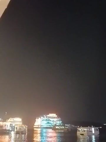 At night, the camera captures a body of water with several illuminated boats. The central boat, larger than the others, features the word “MAJESTIC” in bright lights on its top level. The smaller boats are scattered around it, seemingly docked or slowly moving. The water reflects the lights, creating shimmering patterns. The background is dark, suggesting a night scene.