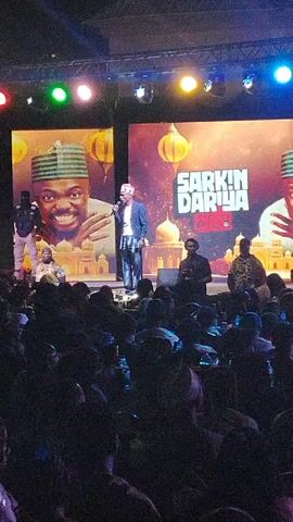 A man in a dark suit and cap is speaking into a microphone on a stage in front of a large audience. Behind him are two large screens, each displaying a picture of the same man with a smiling face, also wearing a cap. The words "Sarkin Dariya" are displayed on the screen to the right. Three other men stand behind the speaker. The stage is lit with various colored lights. The audience is mostly dark, with heads and shoulders visible in the foreground.