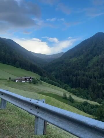 The video pans across a lush green mountainous landscape under a blue sky with scattered clouds. In the foreground, a metal guardrail runs along the bottom of the frame. The middle ground reveals rolling hills covered in dense evergreen forests and a few patches of mowed green fields. A small building with a red roof is visible nestled on one of the hillsides. Further in the distance, the mountains recede, becoming softer and more muted in color, with a small village or town appearing in a valley. The scene suggests a rural, scenic environment, possibly in the Alps, during the daytime.