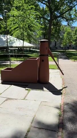 The video opens with a close-up of a reddish-brown water pump with a wide base and a long spout. The pump is situated on a paved area made of large, square stones. To the left of the pump is a matching reddish-brown structure with an angled, stepped design and a basin at the bottom. Behind the pump and structure, a grassy area is visible, bordered by a path and a metal railing. In the background, trees with green leaves provide shade, and beyond them, white tents and a building are partially visible. The lighting suggests it is daytime, with sunlight casting shadows. The camera pans slowly to the left, focusing on the stepped structure and then moving further to reveal more of the grassy park-like environment with trees and buildings.